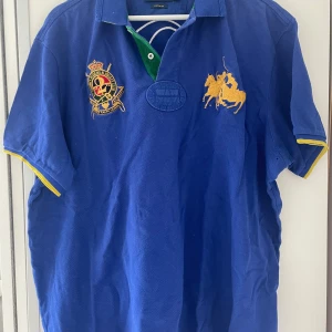 Blå pikétröja Polo Ralph Lauren XXL - Blå pikétröja från Polo Ralph Lauren i bomull, med broderade detaljer och stor gul polospelare på bröstet. Nummer 2 i vitt på ryggen, grönt tyg på insidan av kragen och gula detaljer vid ärmsluten. Custom fit-modell, perfekt för en sportig look.