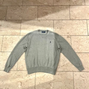 Grå stickad tröja från Polo Ralph Lauren - Snygg grå stickad tröja från Polo Ralph Lauren med klassisk rund hals och broderad logga på bröstet. Tröjan har ribbade muddar vid ärmslut och nederkant, och är långärmad. Perfekt för dig som gillar stilrena och tidlösa plagg. Om ni har några frågor är det bara att höra av sig. Skickar samma dag eller dagen därpå som senast!