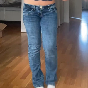Blå bootcut jeans med låg midja - Säljer ett par klassiska blå jeans med bootcut-snitt och låg midja. Jeansen har fem fickor, snygga slitningar och är tillverkade i ett mjukt denimtyg. Perfekta för dig som gillar en avslappnad och tidlös stil. W27 L32