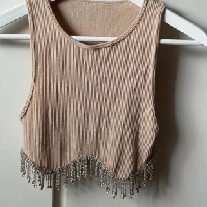 Beige croppad linnetopp med stenar - Säljer en beige croppad linnetopp i ribbad bomull med ärmlös design. Toppen har en vågig nederkant dekorerad med glittrande stenar och fransar i silver som ger extra bling. Tyvärr har den blivit lite nopprig och tappat en Sten, annars är den jättefin!😊