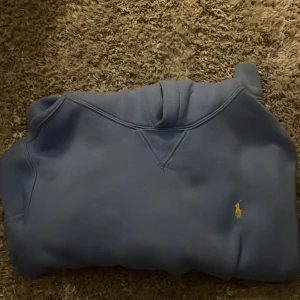 Blå hoodie från Polo Ralph Lauren - Snygg blå hoodie från Polo Ralph Lauren med klassisk gul broderad logga på bröstet. Tröjan har huva och magficka, perfekt för chill dagar. Mjuk insida och stilren design som funkar året runt.