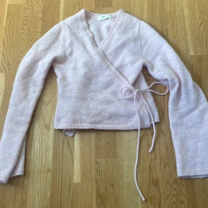 Ljusrosa omlottkofta från Gina Tricot - Supermjuk ljusrosa omlottkofta från Gina Tricot i ullblandning. Koftan har långa ärmar, v-ringning och knytband i sidan. Perfekt för lager-på-lager och ger en söt och chill vibe till din outfit.