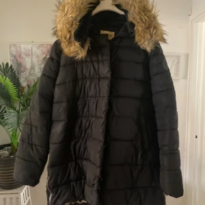 Svart pufferjacka med fuskpäls från Roco Baroco - Svart pufferjacka från Roco Baroco med fuskpäls på huvan och leopardmönstrat innerfoder. Jackan har långa ärmar, dragkedja i sidan och quiltad design. Perfekt för kalla dagar när du vill sticka ut lite extra.