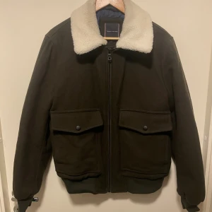 Tommy hilfiger bomberjacka - Snygg mörkbrun bomberjacka från Tommy Hilfiger med vit teddykrage. Jackan har två stora fickor med lock och knappar, och dragkedja framtill. Perfekt för dig som gillar mer klassisk stil