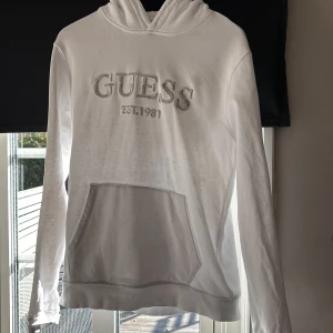 Vit hoodie från Guess med broderad logga - Vit hoodie från Guess med broderad logga och texten 'EST. 1981' framtill. Klassisk känguruficka och huva. Långärmad och i mjukt bomullsmaterial, perfekt för chill dagar. Enkel och stilren design som funkar till det mesta. Den har en fläck på bröstet vet inte var den kommer ifrån, PRIS EJ HUGGET I STEN kom med bud!