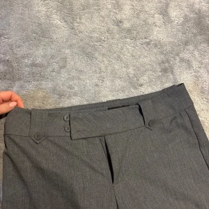 Grå ”Kittie” kostymbyxor från Tiger Mist - Snygga grå lågmidjade kostymbyxor från Tiger Mist med unika knapplösningar i midjan och bakficka med lock. Byxorna har raka ben och är tillverkade i ett mjukt material som ger en clean och stilren look. Det är moderna detaljer på ett klassiskt plagg.