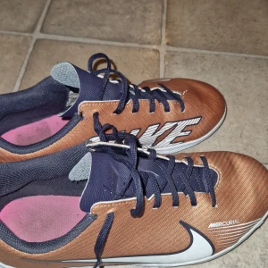 Nike Mercurial fotbollsskor brons - Säljer ett par Nike Mercurial fotbollsskor i brons och marinblått med stora vita loggor på sidorna. Skorna har snörning, platt vit sula och är tillverkade i syntetmaterial. Perfekta för inomhusfotboll och har en riktigt sportig vibe.