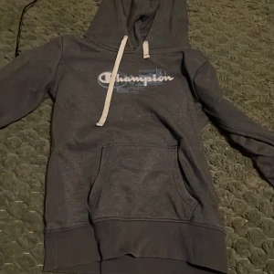 Mörkgrå Champion hoodie med logga - Säljer en mörkgrå hoodie från Champion med stor vit och blå logga på bröstet. Tröjan har luva med vita snören och en stor magficka. Perfekt för chill dagar och streetstyle. Klassisk passform och mjukt material.