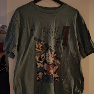 Grön Uniqlo x MFA t-shirt med samurajtryck - Cool grön t-shirt från Uniqlo x Museum of Fine Arts Boston med japanskt samurajmotiv och tryck i flera färger på framsidan. Klassisk passform och rund hals. Perfekt för dig som gillar streetwear och unika prints.