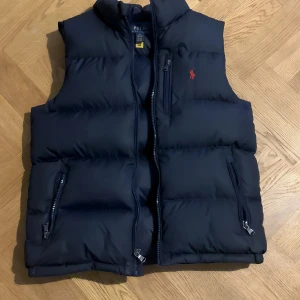 Mörkblå dunväst från Polo Ralph Lauren - Snygg mörkblå dunväst från Polo Ralph Lauren med klassisk röd logga på bröstet. Västen har två dragkedjefickor och en hög krage. Perfekt för lager-på-lager och enkel att matcha med hoodies eller tröjor.