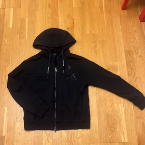 Svart hoodie från Armani Exchange - Svart hoodie från Armani Exchange med dragkedja och huva. Snygga snören med logga och diskret AX-tryck på bröstet. Mjuk bomullskänsla, ribbade muddar och två fickor framtill. Perfekt för chill eller streetwear.