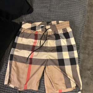 Burberry rutiga badshorts M - Säljer ett par klassiska Burberry badshorts i beige, svart, röd och vit med ikoniskt rutigt mönster. Shortsen har elastisk midja med snörning och en bakficka med dragkedja. Perfekta för sommaren och strandhäng men även vardagligt bruk. Vid frågor är det bara att höra av sig privat!