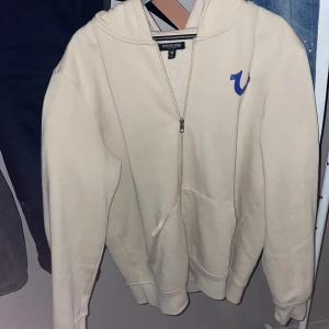 Beige hoodie från True Religion L nypris= 1350!  - Säljer en beige hoodie från True Religion i storlek L. Tröjan har är lite sönder men går enkelt att fixa med tråd och nål!, den har dragkedja, huva och fickor framtill. På bröstet finns True Religion-loggan i blått och på ryggen ett stort tryck med Buddha som spelar gitarr. Perfekt för chill och streetwear.