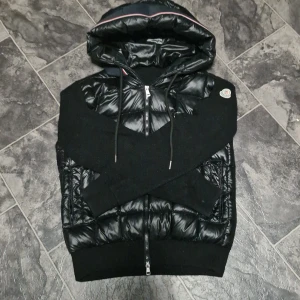 Moncler Jacka - Snygg svart pufferjacka från Moncler med stickade ärmar och quiltade partier i glansigt material. Jackan har huva med dragsko och hel dragkedja framtill. Klassisk Moncler-logga på ärmen och cool detalj med rödvit rand på huvan. Perfekt för både hösten och vintern