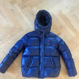 Ralph Lauren vinter jacka - Snygg marinblå pufferjacka från Polo Ralph Lauren med huva och dragkedja. Jackan har det klassiska röda Polo-loggan broderad på bröstet och två fickor framtill. Perfekt för kalla dagar och riktigt skön att bära. Den är i XL teenager men motsvarar S/XS i dam.