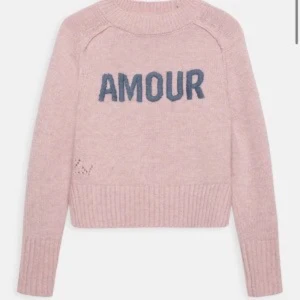 Rosa stickad tröja Zadig & Voltaire - Superfin rosa stickad tröja från Zadig & Voltaire med blå 'AMOUR'-text framtill. Tröjan har ribbade muddar, högre halsringning och diskret hålmönsterdetalj vid höften. Perfekt för dig som gillar en chill men ändå snygg vibe.