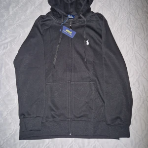 Svart hoodie från Polo Ralph Lauren - Svart hoodie från Polo Ralph Lauren med dragkedja, snörning i huvan och klassisk logga broderad på bröstet. Tröjan har ribbade muddar, fickor framtill och en skön loose passform. Perfekt för chill dagar och streetwear.