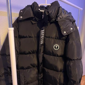 Svart pufferjacka från Trapstar - Säljer en svart pufferjacka från Trapstar med broderad logga på bröstet och stor Trapstar-text på ryggen. Jackan har avtagbar huva, dragkedja och stora fickor framtill. Perfekt för kalla dagar och riktigt streetwear-vibe. Skick 10/10, inga defekter, pris går att diskutera 👌🏼🤝❄️