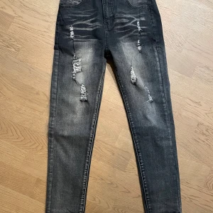 Svarta  jeans med slitningar - Snygga svarta  jeans med slitna detaljer och trasiga partier på låren och knäna. Jeansen har en tvättad look med ljusare partier och klassisk femficksdesign. Perfekta för en trendig och avslappnad stil.