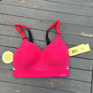 Rosa sport-bh från Triumph Triaction - Sport-bh från Triumph Triaction i stark rosa färg med svarta och röda justerbara axelband. Bh:n har vadderade kupor, meshdetaljer och hakknäppning bak. Snygga silvriga mönster på sidorna och bred resår för extra stöd. Perfekt för träning och cardio.
