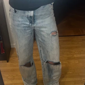 Bershka 90's wide jeans med slitningar - Säljer ett par ljusblå 90's-inspirerade jeans från Bershka i modell wide. Jeansen har hög midja, raka ben och slitna detaljer med hål på båda knäna. Klassisk femficksmodell i denim med cool tvätt och avslappnad vibe.