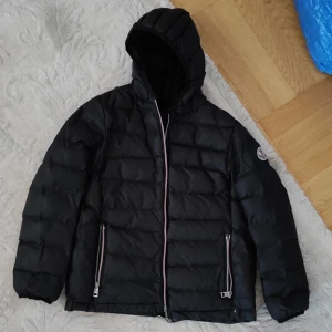 Svart Moncler dunjacka med huva - Svart dunjacka från Moncler med huva och klassisk logga på ärmen. Jackan har quiltad design, två fickor med dragkedja och snyggt broderad Moncler-text på ryggen. Perfekt för kalla dagar och riktigt stilren look. Har många hål men inget som syns ne man har på sig den alla är inne i jackan och dragkedja är sönder går att fixa 