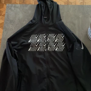 Svart vindjacka med Nike-tryck - Svart vindjacka från Nike med huva och hel dragkedja. Framsidan har ett stort vitt Nike-tryck med swoosh-loggor och text. Jackan är tillverkad i polyester och har långa ärmar med diskret Nike-logga på ena ärmen. Perfekt för en sportig och stilren look.