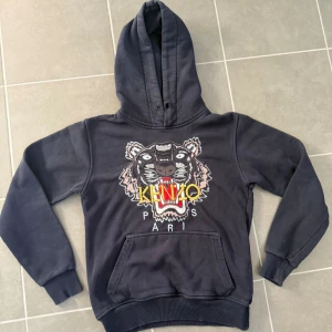 Mörkblå Kenzo hoodie med tigerbrodyr - Mörkblå hoodie från Kenzo med ikonisk broderad tiger och färgglad logga på bröstet. Tröjan har huva, magficka och ribbade muddar. Perfekt för dig som vill sticka ut med en streetstyle-look.