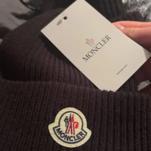 Svart ribbad mössa från Moncler - Säljer en svart ribbstickad mössa från Moncler med klassisk uppvikt kant och broderad logotyp framtill. Perfekt accessoar för kalla dagar och snygg till alla outfits.