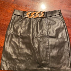 Svart skinnkjol med kedjedetalj Amisu - Snygg svart kort skinnkjol från Amisu med en stor guldfärgad kedjedetalj i midjan. Kjolen har en figurnära passform och dragkedja bak. Perfekt för dig som vill ha en edgy och trendig look.