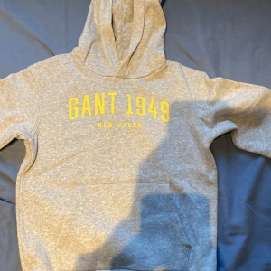 Grå hoodie från Gant med tryck - Snygg grå hoodie från Gant med gult tryck 'GANT 1949 NEW HAVEN' på bröstet. Klassisk känguruficka framtill, huva och långa ärmar med muddar. Perfekt för chill dagar eller när du vill ha en sportig look.