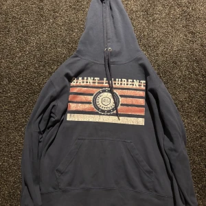 Saint Laurent marinblå hoodie med tryck - Säljer en marinblå hoodie från Saint Laurent med stort tryck framtill i rött, vitt och blått. Tröjan har huva med snörning, känguruficka och långa ärmar. Perfekt för dig som vill ha en snygg och bekväm streetstyle-look.