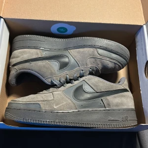 Grå Nike Air Force 1 sneakers mocka - Säljer ett par grå Nike Air Force 1 sneakers i mocka med mörkgrå detaljer och svart swoosh. Skorna har klassisk låg siluett, rund tå och snörning. Snygg platt sula och stilren look som passar till streetwear. Perfekt för dig som gillar neutrala färger och ikoniska modeller. Kom med bud.
