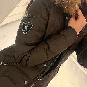 Svart dunjacka Rockandblue med päls - Snygg svart dunjacka från Rockandblue med fluffig päls på kragen. Jackan har quiltad design, dragkedja och knappar framtill samt en patch på ärmen. Perfekt för kalla dagar och har en cool, urban vibe. Pälsen är äkta tvättbjörn, köptes för cirka 4000kr.