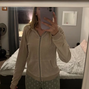 Hoodie - Mysig beige hoodie i mjuk velour med dragkedja framtill och huva. Tröjan har långa ärmar, ribbade muddar och två fickor framtill. Perfekt för chill dagar eller när du vill vara extra bekväm.