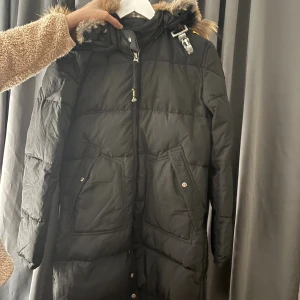 Svart Parajumpers lång dunjacka - Svart lång dunjacka från Parajumpers med gul detalj på axeln, patch på ärmen och fuskpäls runt huvan. Jackan har dragkedja, knappar och stora fickor framtill. Perfekt för kalla dagar med sin quiltade design och snygga look.