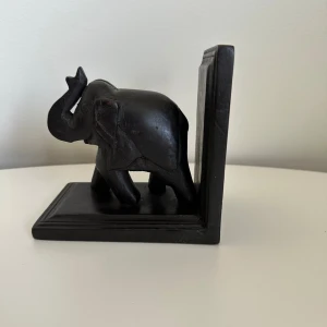 Bokstöd i trä - Ett dekorativt bokstöd i trä, format som en elefant. Perfekt för att hålla ordning på dina böcker med en touch av elegans. Mycket gott skick.   Hämtas i Stockholm eller skickas mot porto. (Boken medföljer ej)