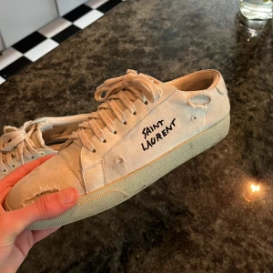 Beige Saint laurent skor med sliten look - Saint Laurent sneakers i beige canvas med handskriven logga på sidan och slitna detaljer för en rå streetstil. Skorna har snörning, rund tå och platt sula. Baksidan har präglad Saint Laurent-text och inslag av skinn. Perfekt för dig som gillar designer. Sköna skor som passar till allt.