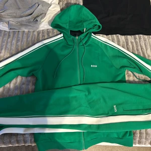 Grön traucksuit från BOSS  - Snygg grön traucksuit från BOSS med vita ränder längs sidorna och elastisk midja. Traucksuiten har en sportig vibe och är tillverkad i mjuk bomullsmix. Perfekta för chill eller träning, med diskret BOSS-logga på benet. Använt endast 1 gång vill sälja då det var en present men var för stor.