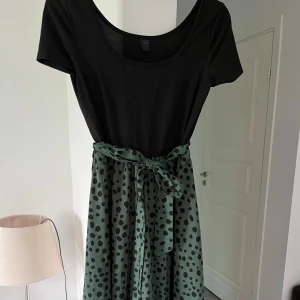 Svart och grön klänning med knytband - Supersnygg klänning med svart ribbad överdel och grön kjol med svart prickigt mönster. Kjolen har ett matchande knytband i midjan som ger en extra detalj. Kortärmad och med rund halsringning, perfekt för dig som gillar en cool och avslappnad stil.