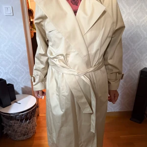 Beige trenchcoat med bälte - Snygg beige trenchcoat med klassisk omlott-design och bälte i midjan. Jackan har långa ärmar med spännen vid handlederna och bred krage. Perfekt för dig som vill ha en stilren och tidlös look.