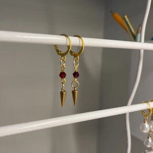 Guldiga örhängen med röda pärlor - Snygga guldiga örhängen med små röda pärlor och spetsiga konformade hängen. Perfekt för dig som gillar att sticka ut med coola detaljer och färg. Passar till både street och trendiga looks. I rostfritt stål och handgjort