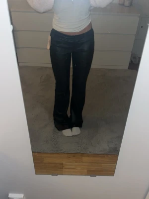 Svarta PU flare-byxor från Nelly (helt oanvända) - Snygga svarta byxor från Nelly i PU-material med låg midja och utsvängda ben. Byxorna har en stilren look med knapp och dragkedja framtill. Perfekt för dig som vill ha en trendig och edgy vibe i garderoben. Helt nya och oanvända och helt slutsålda överallt! 
