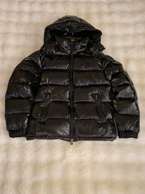 Svart pufferjacka från Moncler - Säljer en svart pufferjacka från Moncler med glansig finish och huva. Jackan har två framfickor med dragkedja, elastiska muddar och klassisk Moncler-logga på ärmen. Perfekt för kalla dagar och har en riktigt snygg streetstil.