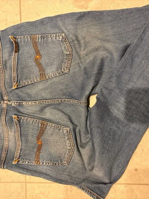 Nudie Jeans blå jeans W31 L32 - Säljer ett par klassiska blå jeans från Nudie Jeans med snygga orangea sömmar och ikonisk brodering på bakfickorna. Modellen har normal passform och raka ben, tillverkade i slitstarkt bomullsmaterial. Perfekt för dig som gillar stilrena och tidlösa jeans.