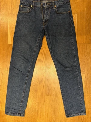 Woodbird jeans i mörkblå denim - Säljer ett par mörkblå jeans från Woodbird style: DOC 90s i tvätten 90s blue. Klassisk femficksdesign, raka ben och normal passform. Orangea sömmar och läderpatch bak i midjan. Perfekt för dig som gillar stilrena och tidlösa jeans.