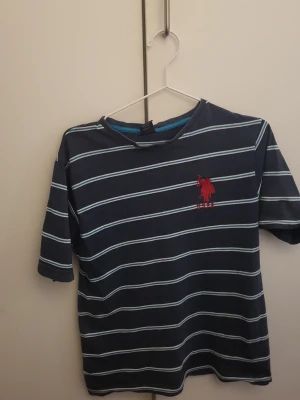 Randig t-shirt från USPA, POLO, marinblå - Säljer en marinblå t-shirt från USPA med vita och ljusblå ränder. T-shirten har korta ärmar och ett rött broderat USPA-logotyp på bröstet. Materialet är mjuk bomull och passformen är normal. Perfekt för dig som gillar klassisk stil med en sportig touch.