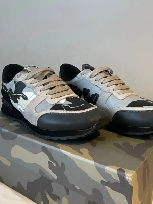 Valentino Garavani Rockrunner  - Säljer ett par Valentino Garavani Rockrunner sneakers i storlek 43. Skorna har ett snyggt camouflage-mönster i svart, grått och beige med detaljer i mocka och skinn. Platt sula med mönstrad undersida och snörning framtill. Märkeslogga på plösen och box medföljer.