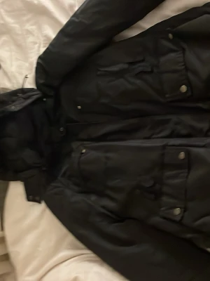 Svart parkajacka från Brema, strl 48 - Svart parkajacka från Brema med flera fickor framtill och snygga tryckknappar. Jackan har huva och är tillverkad i slitstark polyester, perfekt för kyliga dagar. Designad i Italien och har en klassisk, lite loose passform.