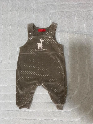 Brun prickig byxdress med rådjur - Söt brun byxdress med små vita prickar och ett broderat rådjur på bröstet. Byxdressen har axelband med knappar och tryckknappar vid benen för enkel på- och avklädning. Mjuk och skön kvalitet, perfekt för chill dagar.